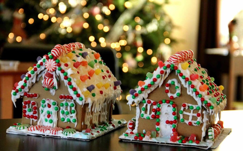 2 gingerbread houses decorated with colorful candies & icing on background of blurred Christmas tree with lights - FAS Exteriors