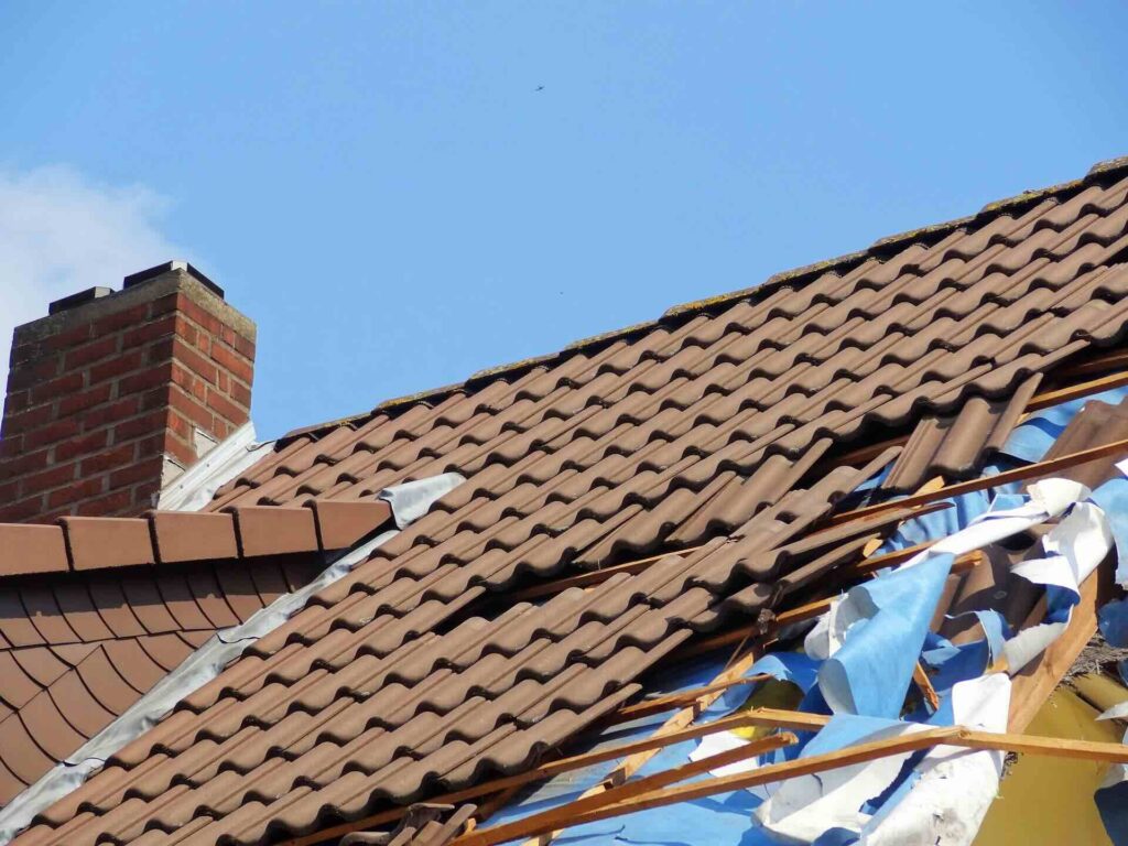 Storm-damaged, brown tiled roof, torn tiles exposing blue underlayment and wooden battens against clear, blue sky - FAS Exteriors