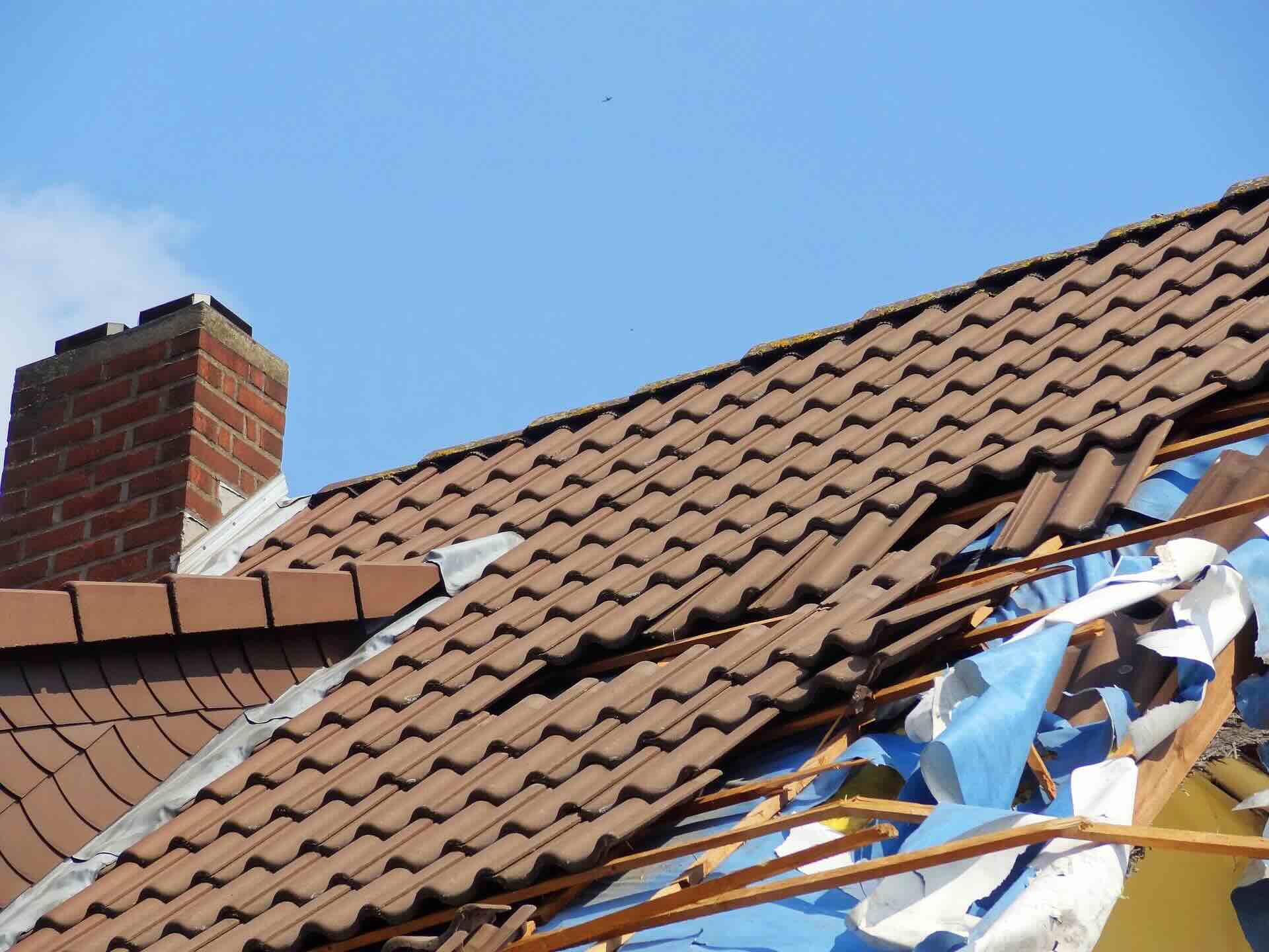 Storm-damaged, brown tiled roof, torn tiles exposing blue underlayment and wooden battens against clear, blue sky - FAS Exteriors